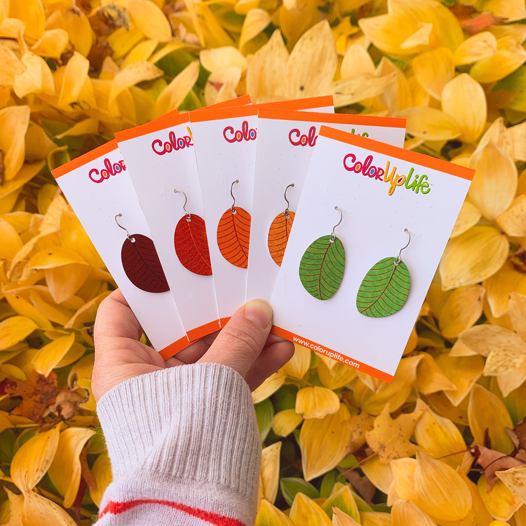 A hand holding five pairs of fall-colored small leaf dangle earrings in front of fall leaves in nature by ColorUpLIfe.