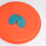 A pair of turquoise small leaf earrings displayed on an orange surface by ColorUpLife.